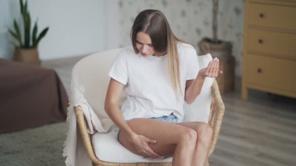 Mujer joven se sienta en el sillón y aplica crema anti-celulitis en su pierna — Vídeos de Stock