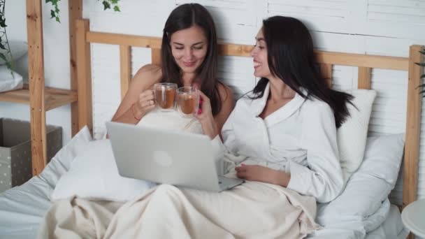 Дві жінки друзі сидять у ліжку, п'ють чай, дивляться смішний фільм на ноутбуці, сміються — стокове відео