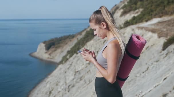 Atleta ragazza in abbigliamento sportivo con tipi di tappetino messaggio di testo sul telefono, mare sullo sfondo — Video Stock