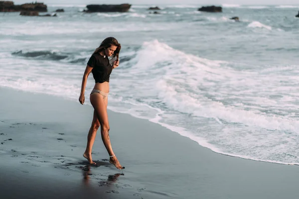 Siyah t-shirt ve mayo seksi vücut ile Kız siyah kumlu plajda yürüyor — Stok fotoğraf