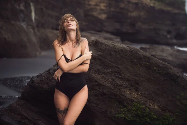 Sexy girl in swimsuit with tattoo on leg rests on black sandy beach on vacation — Stock Photo, Image