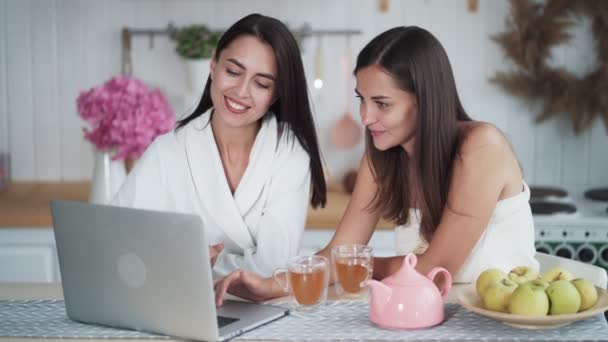 Dos mujeres jóvenes miran algo en el ordenador portátil, discutir, desayunar en la cocina . — Vídeos de Stock