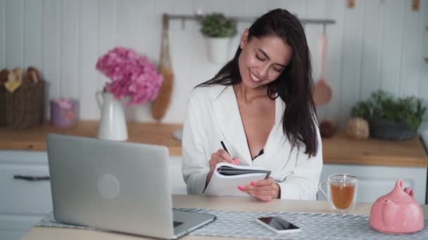 Jeune femme en peignoir assis sur la cuisine et prend des notes dans un carnet — Video