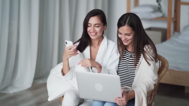 Due donne sedute in poltrona in camera da letto tengono laptop, telefono, card e negozio online — Video Stock