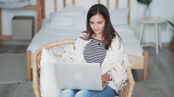 Belle jeune femme est assise dans un fauteuil et utilise un ordinateur portable. Freelancer travaille à la maison — Video