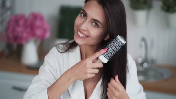 Jeune femme en peignoir blanc peigne ses cheveux avec peigne, regarde la caméra, sourit — Video