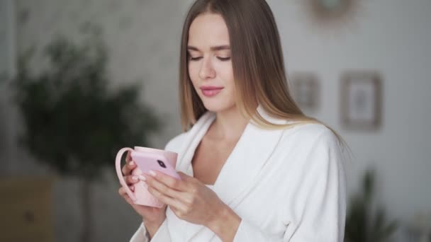 Retrato de mujer rubia joven utiliza el teléfono y sostiene la taza de café o té en la cocina — Vídeo de stock