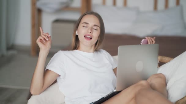 Dívka sedí v slaměném křesle s laptopem, poslouchá hudbu v bezdrátovém sluchátku, tance — Stock video