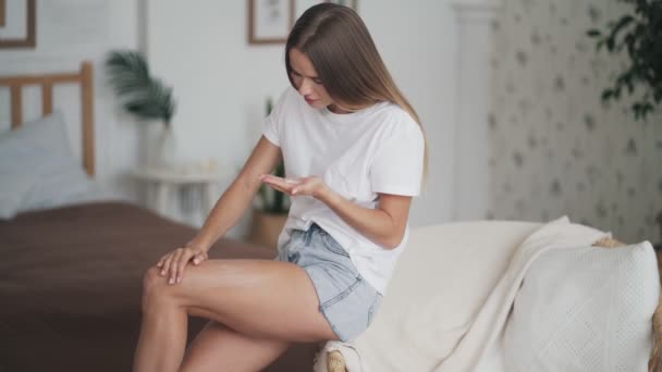 Νεαρή γυναίκα κάθεται στην πολυθρόνα και εφαρμόζει κρέμα κατά της κυτταρίτιδας στο πόδι της — Αρχείο Βίντεο