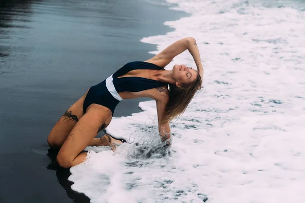 Menina sexy em roupa de banho azul senta-se na praia com areia preta e ondas grandes Fotos De Bancos De Imagens