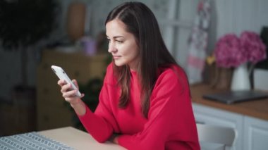 Genç bir kadın mutfakta oturuyor, telefon kullanıyor, internette sohbet ediyor ya da uzaktan çalışıyor.