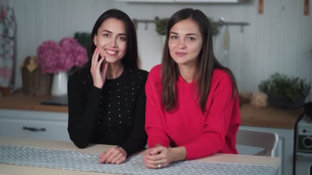 Δύο όμορφα καστανά κορίτσια κάθονται στο τραπέζι στην κουζίνα, κοιτώντας την κάμερα, χαμογελώντας — Αρχείο Βίντεο