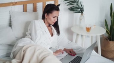 Bornozlu genç kadın yatağa uzanıyor ve çalışmak ya da internetten çalışmak için dizüstü bilgisayar kullanıyor.