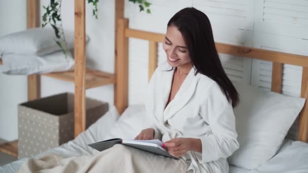 Mladá hezká žena v bílém županu leží na posteli a čte knihu — Stock video