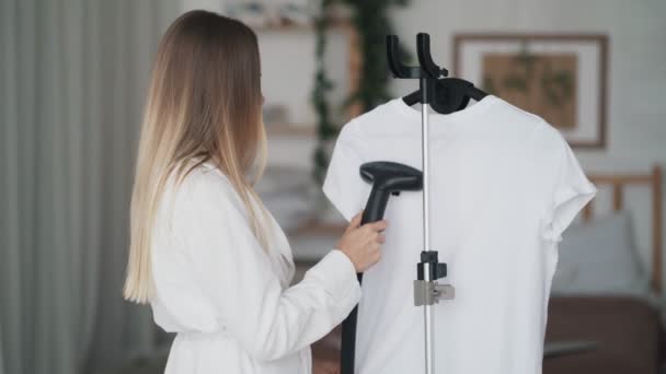 Mooie jonge vrouw in witte badjas met stoomsysteem voor strijk kleding — Stockvideo