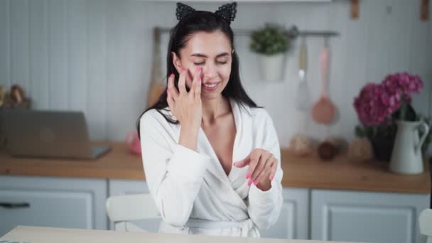 Jovem mulher em roupão aplica creme no rosto e faz massagem facial — Vídeo de Stock