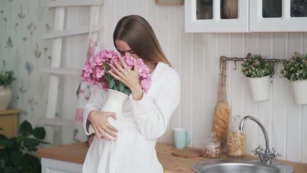 Boldog fiatal nő fehér fürdőköpeny tartja csokor virágot, szippantás azt, mosolyog — Stock videók