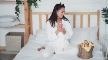 Bornozlu genç kadın yatakta çay içiyor ve yeni pişmiş baget yiyor.