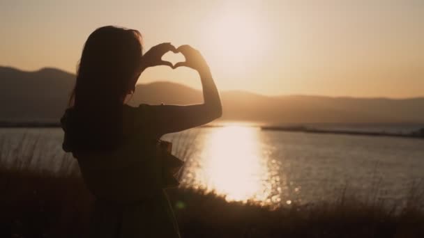 Mãos de mulher faz forma de coração com oceano e pôr do sol no fundo, câmera lenta — Vídeo de Stock