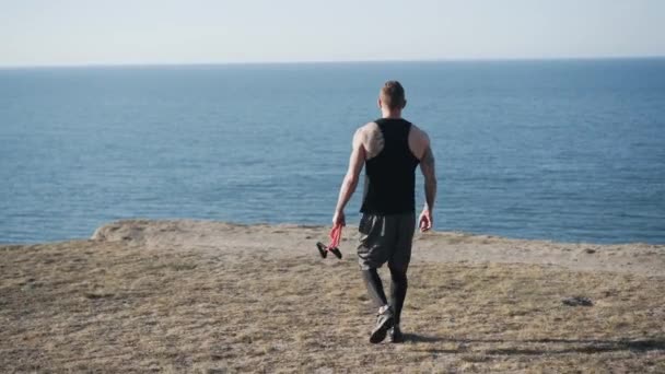 Vue de derrière, l'homme va avec des élastiques de fitness sur la montagne, vue sur la mer — Video