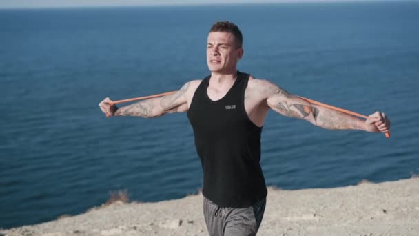 Tetovált sportoló csinál gyakorlatokat rugalmas szalagok óceánra néző — Stock videók