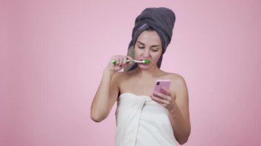 Havlu giyen kız dişlerini fırçalıyor ve pembe arka planda izole edilmiş akıllı telefon kullanıyor.