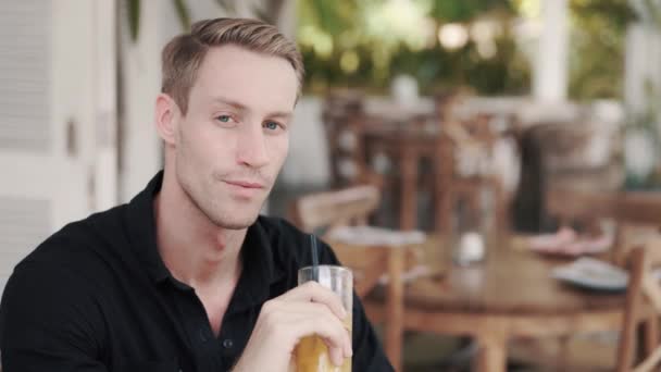 Retrato del hombre rubio bebiendo jugo fresco en la cafetería, mira a la cámara, sonríe — Vídeos de Stock