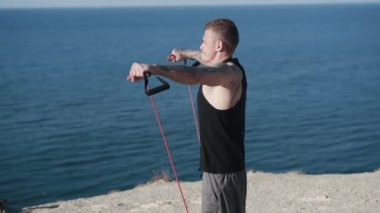 Kas dövmeli adam dışarıda deniz manzaralı fitness elastik bantlarla antrenman yapıyor.