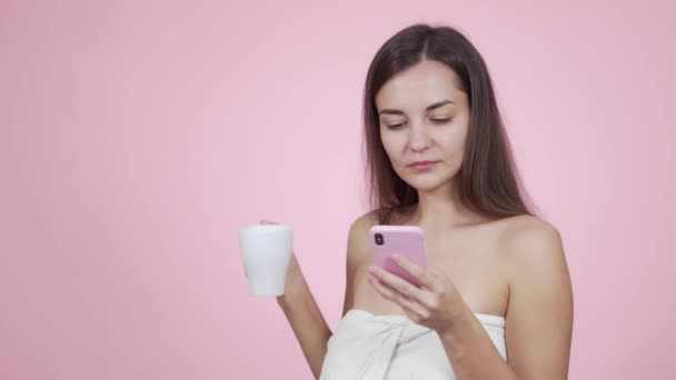 Vrouw in bad handdoek maakt gebruik van smartphone, drinkt koffie geïsoleerd op roze achtergrond — Stockvideo