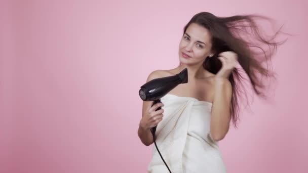 Girl in bath towel dries wet hair with hairdryer isolated on pink background — Stock Video