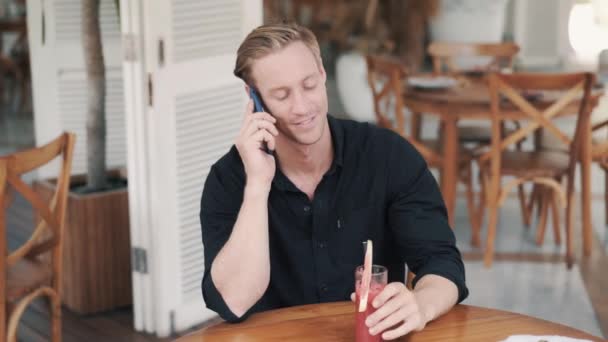 Joven hablando por teléfono, bebiendo jugo de sandía en la cafetería, mirando a la cámara — Vídeos de Stock