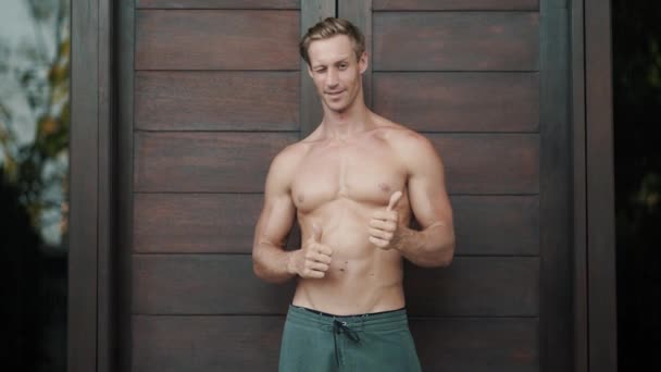 Без рубашки мускулистый человек показывает знак большим пальцем вверх, хорошо, подмигивает, деревянная дверь на заднем плане — стоковое видео