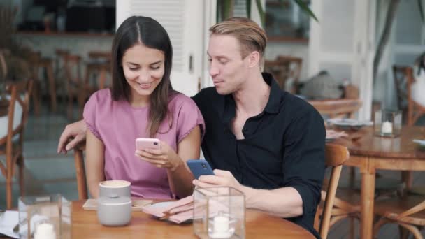 Молодой человек и женщина сидят в кафе, смотрят на смартфон, смеются и разговаривают — стоковое видео