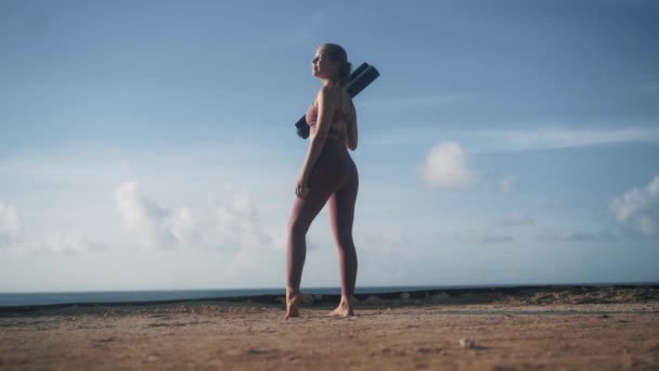 Mujer en ropa deportiva en la playa sostiene estera de yoga, mira a su alrededor, disfruta de la mañana — Vídeo de stock