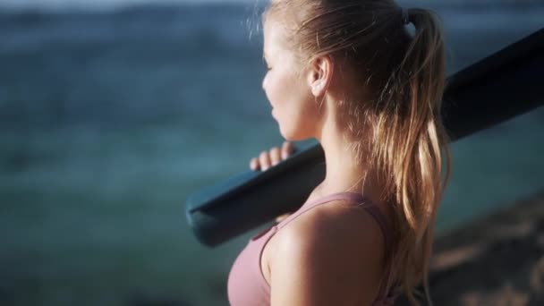Портрет жінки в спортивному одязі на пляжі тримає йога килимок, дивиться на океан — стокове відео