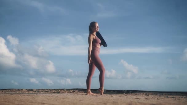 Дівчина в спортивному одязі на пляжі тримає йога килимок, дивиться навколо, насолоджується ранковим часом — стокове відео