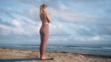 Genç sporcu kadın, yavaş çekimde plajda jimnastik minderi üzerinde yoga egzersizleri yapıyor.
