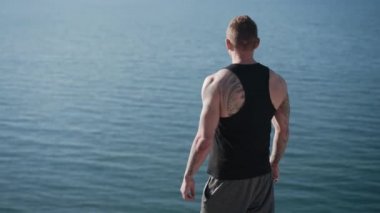 Arka manzara, dövmeli adam dağın tepesinde duruyor, deniz manzarasına bakıyor.