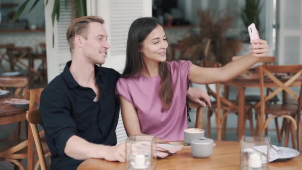 Щасливий хлопець і дівчина сидять у відкритому кафе, п'ють каву і приймають селфі по телефону — стокове відео