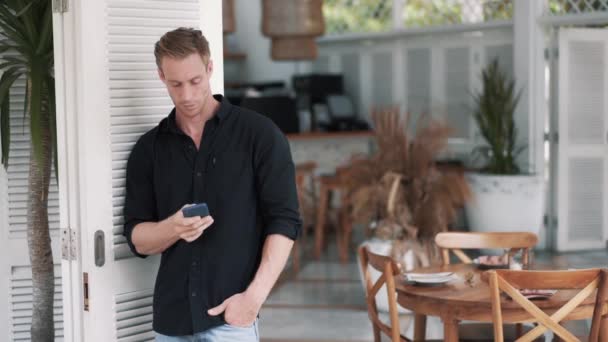 Hombre guapo de camisa negra se para en la cafetería con estilo y utiliza el teléfono, enviar mensaje — Vídeos de Stock