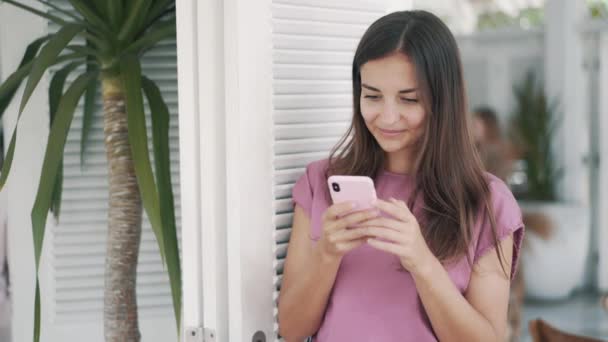 Женщина в кафе на открытом воздухе и с помощью мобильного телефона, улыбается и смотрит в камеру — стоковое видео
