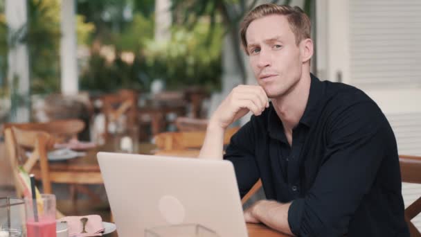 Επιχειρηματίας κάθεται στο τραπέζι με φορητό υπολογιστή στο σύγχρονο καφέ, κοιτάζει κάμερα — Αρχείο Βίντεο