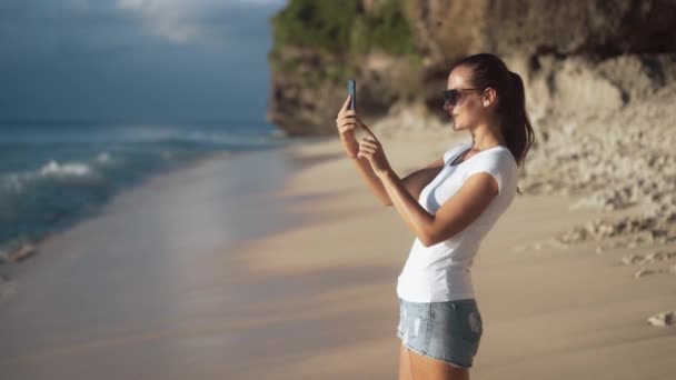 Весела молода красива жінка в сонцезахисних окулярах фотографує пляж — стокове відео