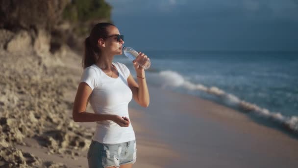 O femeie tânără frumoasă în ochelari de soare bea apă limpede din sticlă pe plajă — Videoclip de stoc