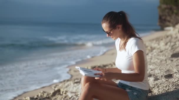 Чарівна молода дівчина читає книгу, сидячи на піщаному березі — стокове відео