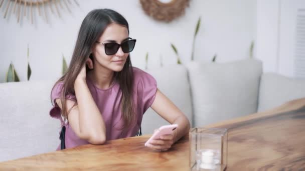 Портрет красивої жінки в темних сонцезахисних окулярах сидить в кафе і використовує телефон — стокове відео