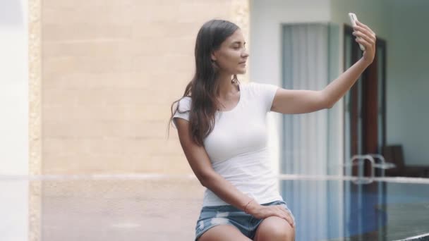 Retrato de chica viajera blogger hace selfie en el teléfono, sentado en el borde de la piscina — Vídeos de Stock