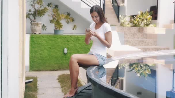Nyári ruhás fiatal nő mobilon ül a medence szélén. — Stock videók