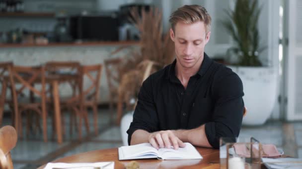 Сосредоточенный мужчина в черной рубашке сидит в современном открытом кафе и читает книгу — стоковое видео