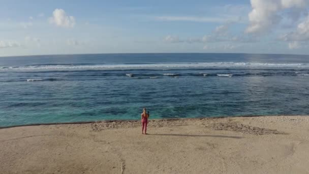 Vue aérienne, jeune femme avec tapis de gymnastique se tient sur la plage et regarde l'océan — Video